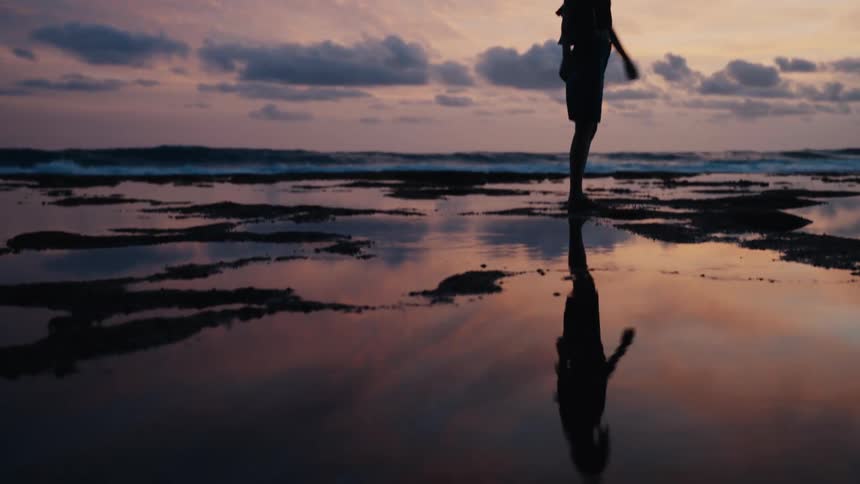 A silhouetted woman walks slowly through reflective tide pools on a beach during a beautiful pastel sunset