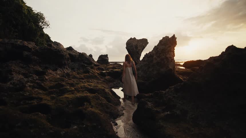 A woman in a white dress walks slowly through a rocky tide pool path on a beautiful beach at sunset