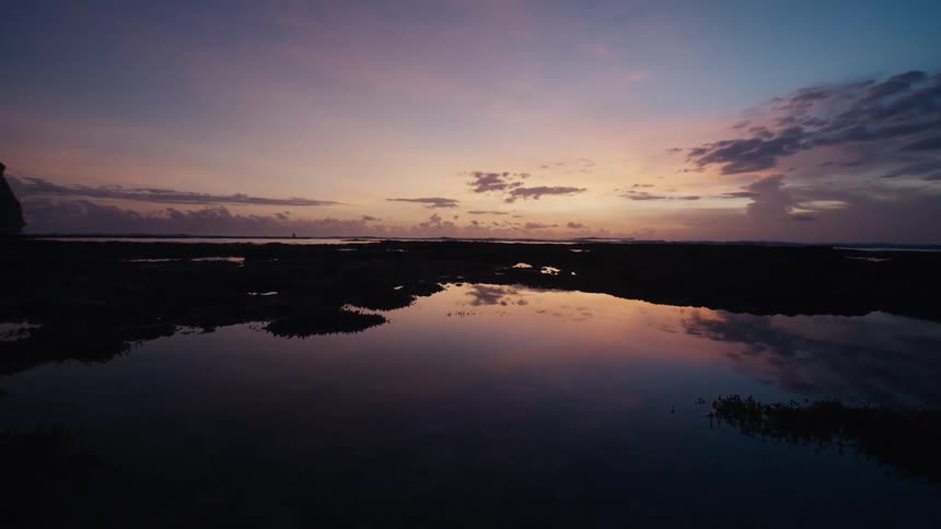 A tranquil coastal landscape at sunset shows a beautiful pink and purple sky perfectly reflecting in the calm tidal pools