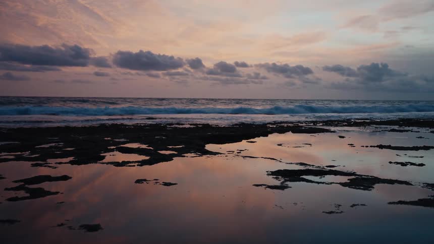 The colorful sunset sky reflects beautifully in the tide pools of a rocky beach as ocean waves crash in the background