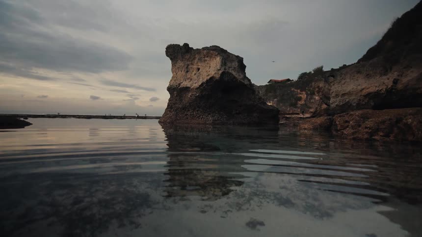 A serene, low-angle view of a tidal pool reflecting a large rock formation under a moody, overcast sky