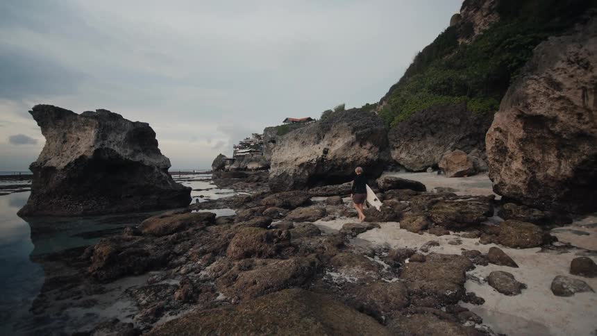 A lone surfer carries their surfboard along a rugged, rocky shoreline at low tide, with distant cliffside buildings under a moody, overcast sky