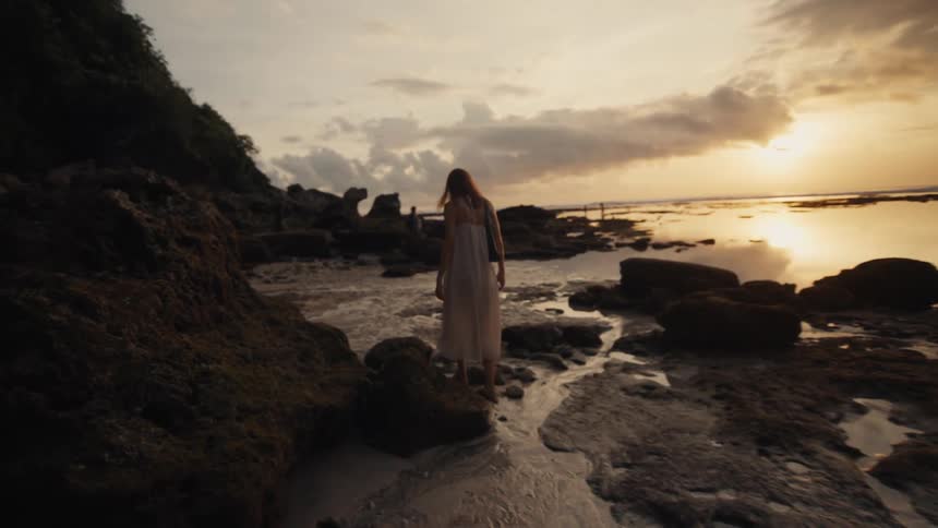 In slow motion, a woman in a white dress gracefully walks along the wet, rocky shoreline as the golden sun sets over the calm ocean