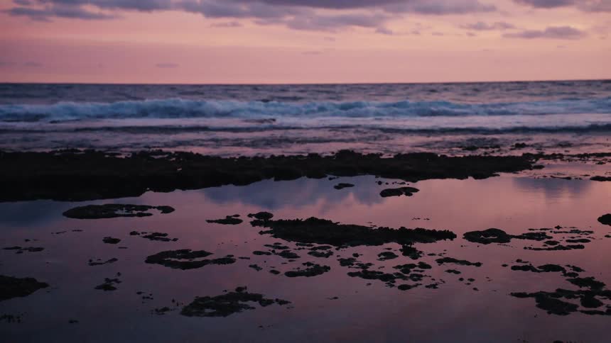 Calm tide pools on a rocky shoreline reflect a stunning pink and purple sunset over the ocean