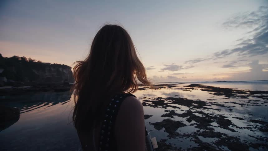 A thoughtful young woman in slow motion watches the calm ocean from a rocky beach as the twilight sky reflects in the water