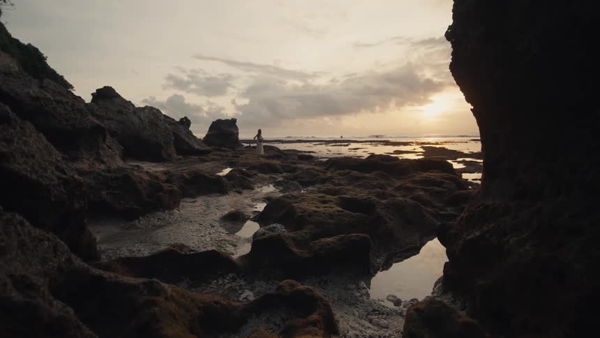 A lone person in a dress stands on a rugged, rocky beach watching the beautiful sunset over the ocean