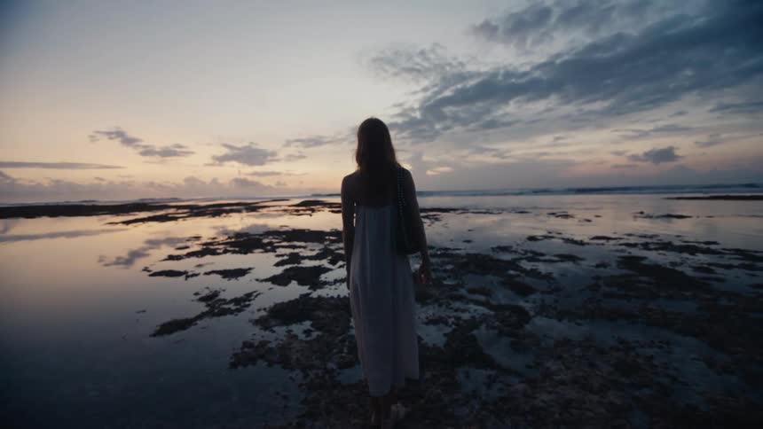 A lone woman in a flowing dress contemplates the serene ocean landscape during a beautiful twilight sunset
