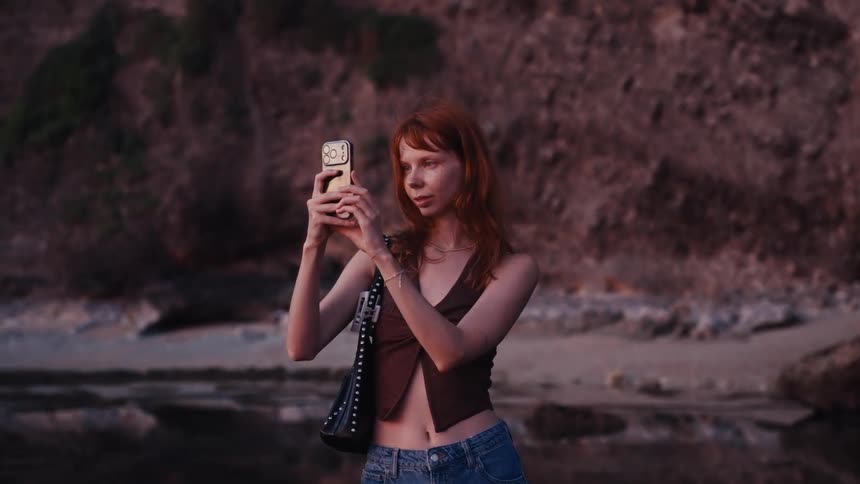 A stylish woman with red hair takes a photo on her smartphone at a rocky beach during a beautiful sunset