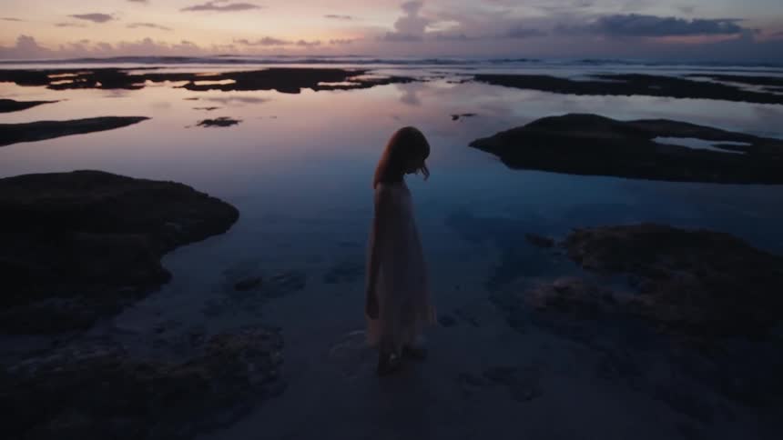 A woman in a long dress is silhouetted as she walks through shallow tide pools on the beach during a beautiful twilight