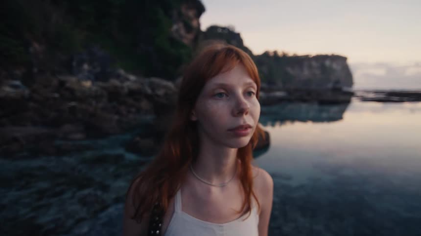 A close-up slow-motion shot of a pensive young woman with red hair and freckles looking out over a calm ocean at sunset