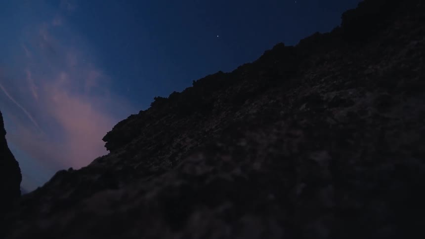 A low-angle time-lapse captures fireflies dancing over a rocky landscape under a starry night sky