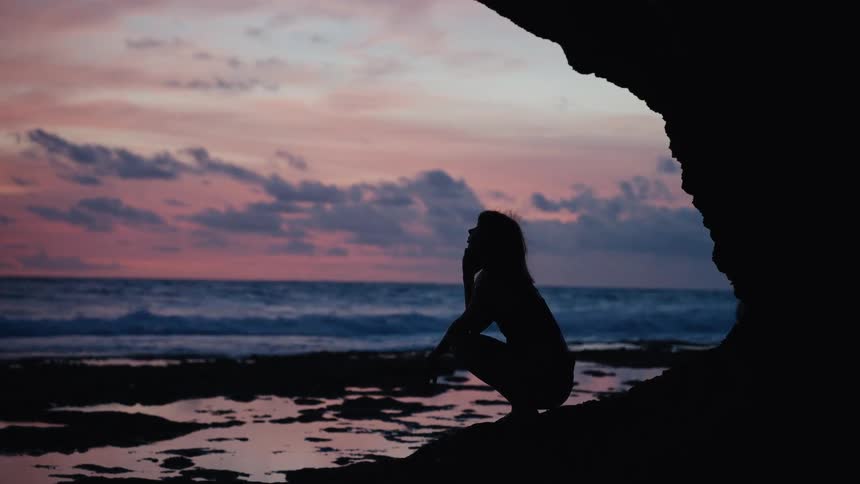 A silhouetted woman crouches pensively in a cave on a rocky beach, watching the waves during a beautiful pink sunset