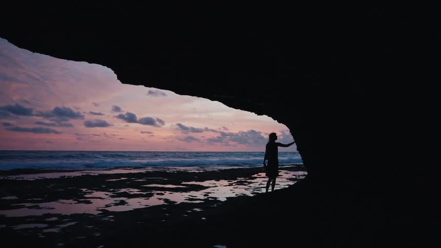 A silhouetted woman walks to the edge of a cave to watch the beautiful pink sunset over the ocean
