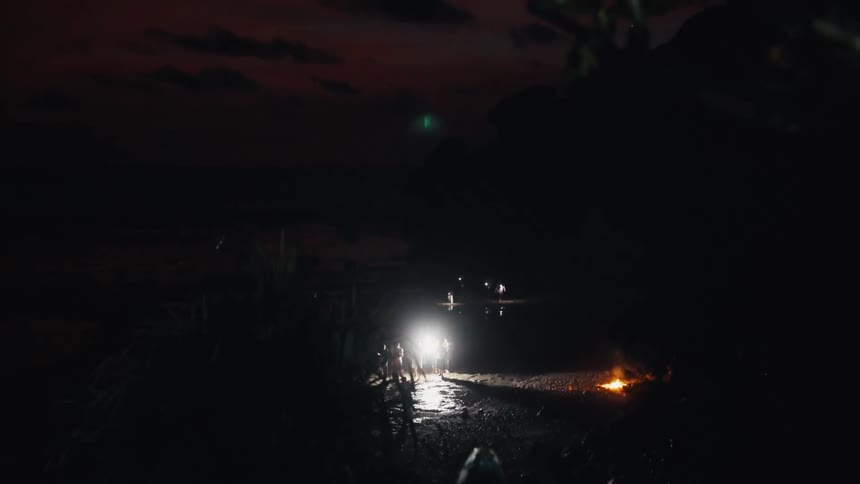 Friends gather on a tropical beach at dusk, their figures silhouetted by the bright headlights of a car and a warm bonfire