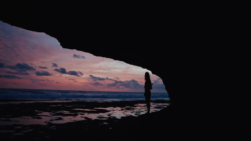 A woman's silhouette stands inside a dark beach cave, peacefully watching the beautiful pink and purple sunset over the ocean waves