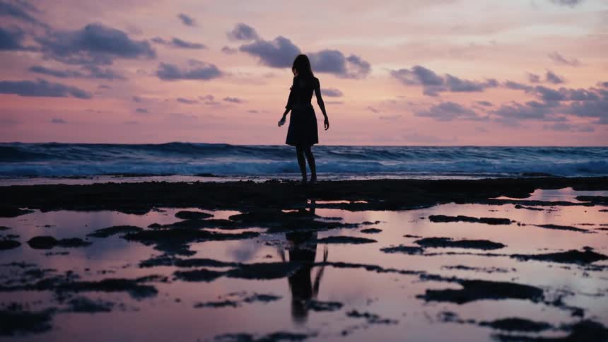 A serene woman in silhouette stretches and walks along a rocky beach against a stunning pink sunset sky