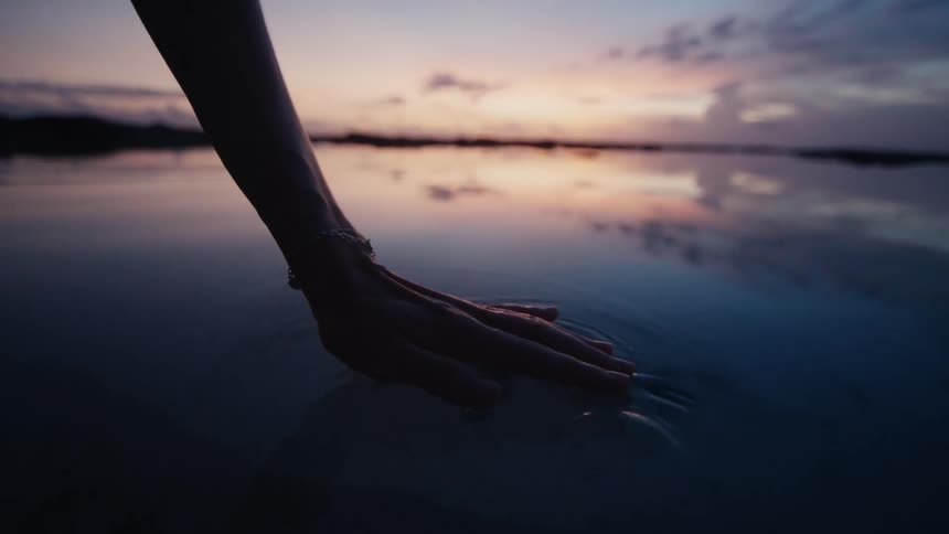 A silhouetted hand gently touches the calm water at sunset, creating ripples in slow motion