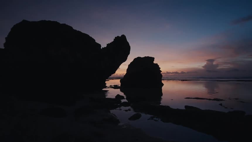 Silhouetted rock formations on a calm beach reflect the colorful sky at twilight