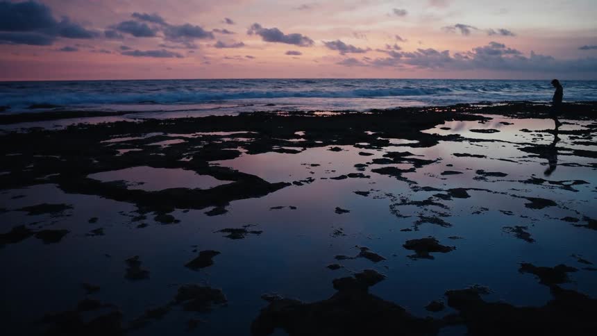 A silhouetted person walks along a rocky beach filled with tide pools that reflect the vibrant colors of the sky at dusk