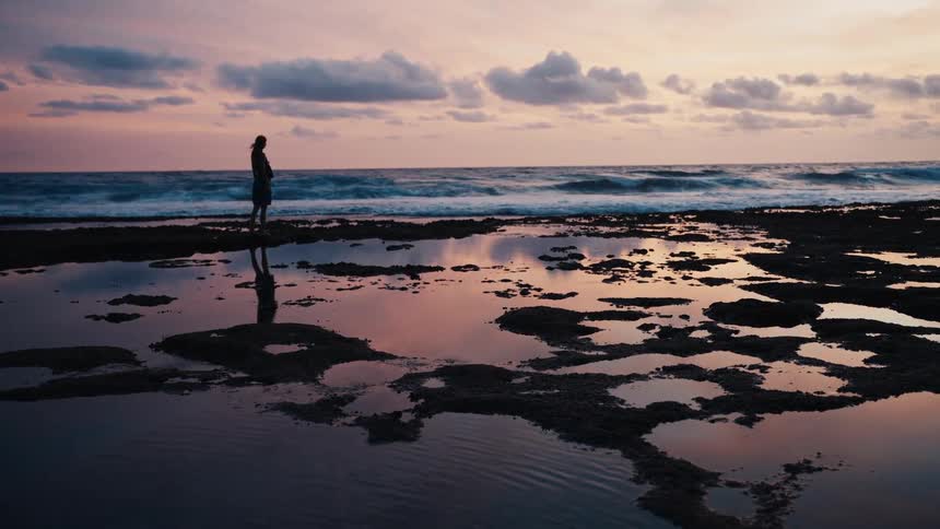 A woman in silhouette walks along a rocky shore with tide pools reflecting the beautiful pastel sunset sky as she stops to watch the ocean