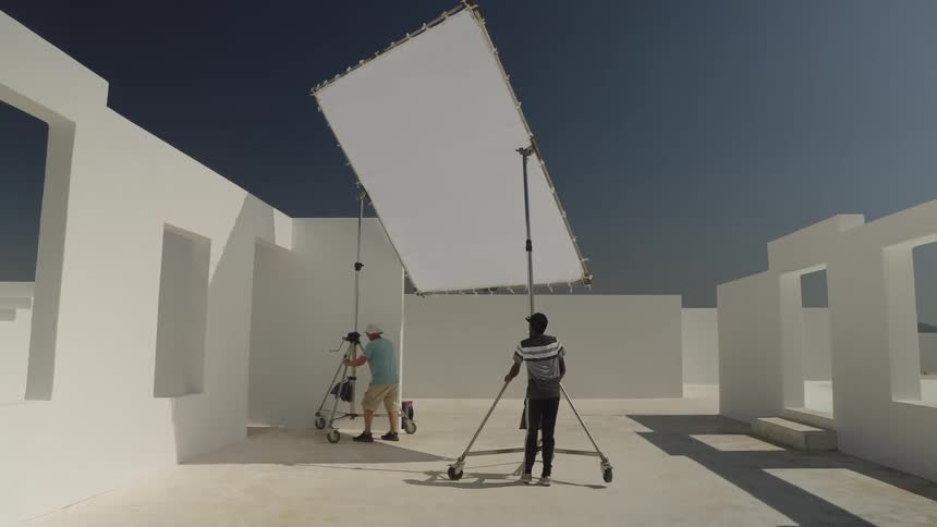 A film crew with a camera and large light diffuser prepares for a shoot on a minimalist white architectural set outdoors
