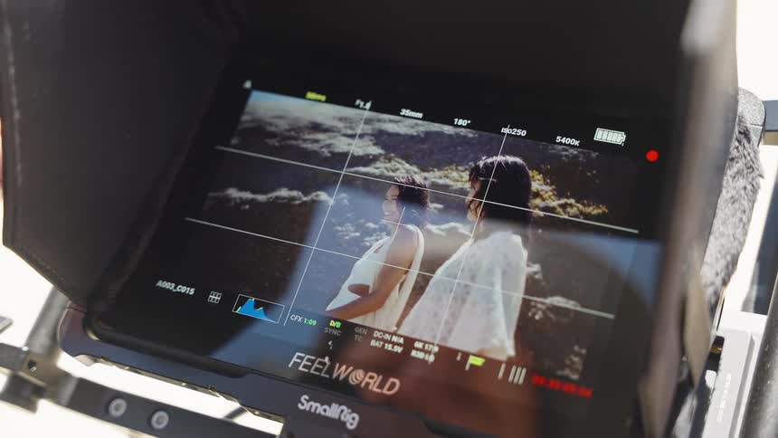 A close-up of a camera monitor displays a scene of two models posing on a sunny beach during a film shoot