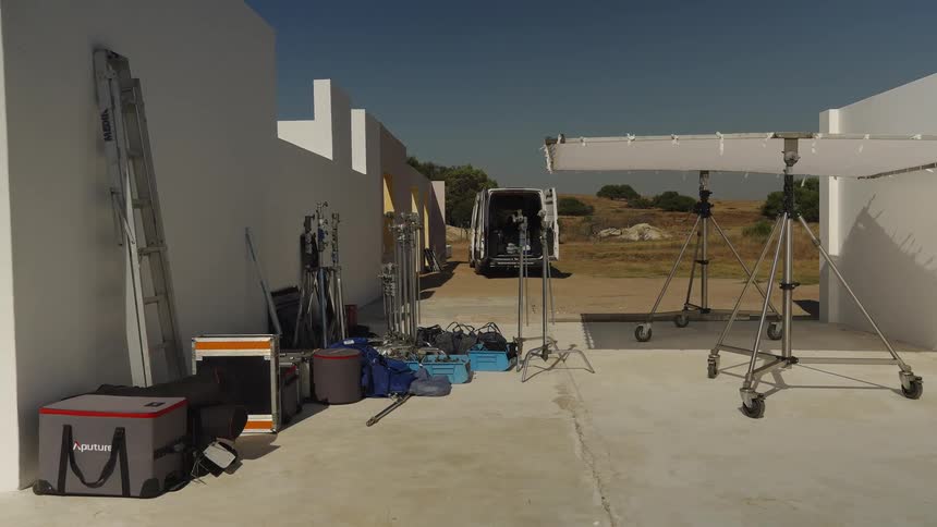 A wide, static shot reveals a film production set with professional lighting equipment arranged between modern white walls under a clear blue sky