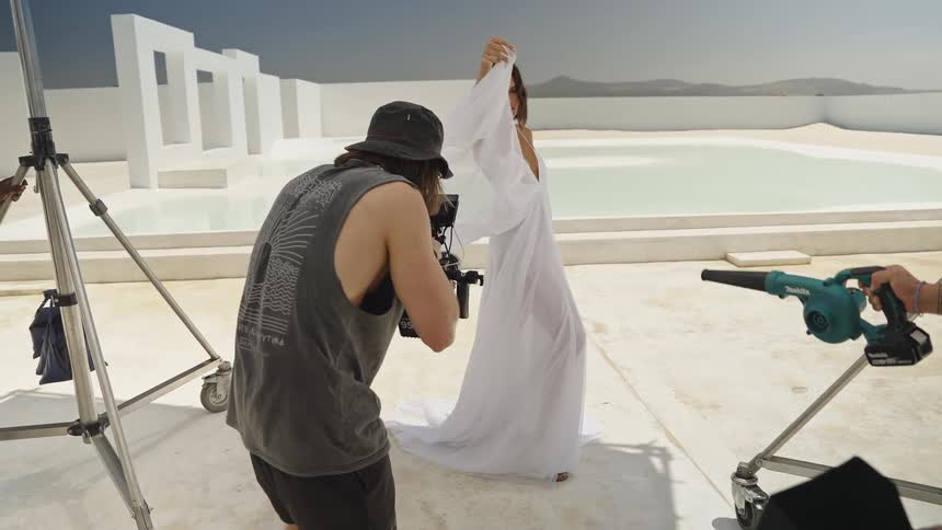 A slow-motion, behind-the-scenes look at a fashion photoshoot with a model in a flowing white dress, a videographer, and an assistant using a wind blower