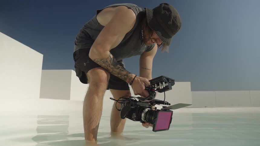 A filmmaker stands barefoot in a shallow white pool under a clear blue sky, carefully setting up a shot with his cinema camera