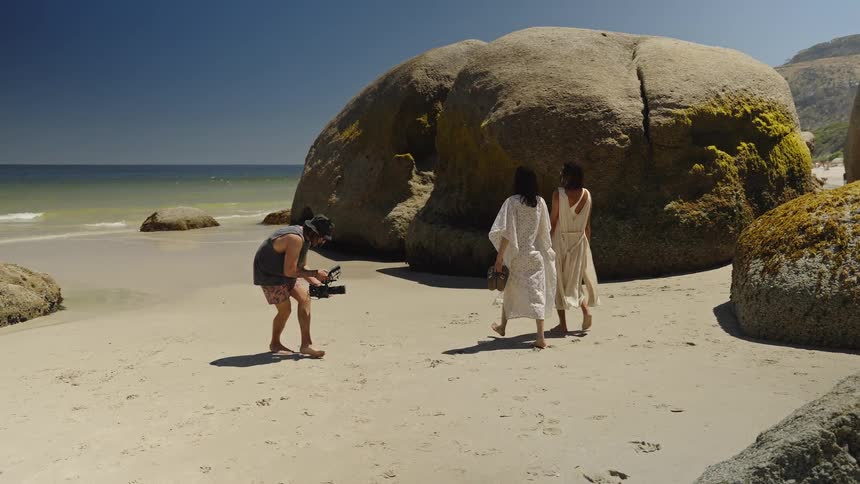 A videographer films two women in flowing white dresses walking barefoot on a sandy beach with large boulders