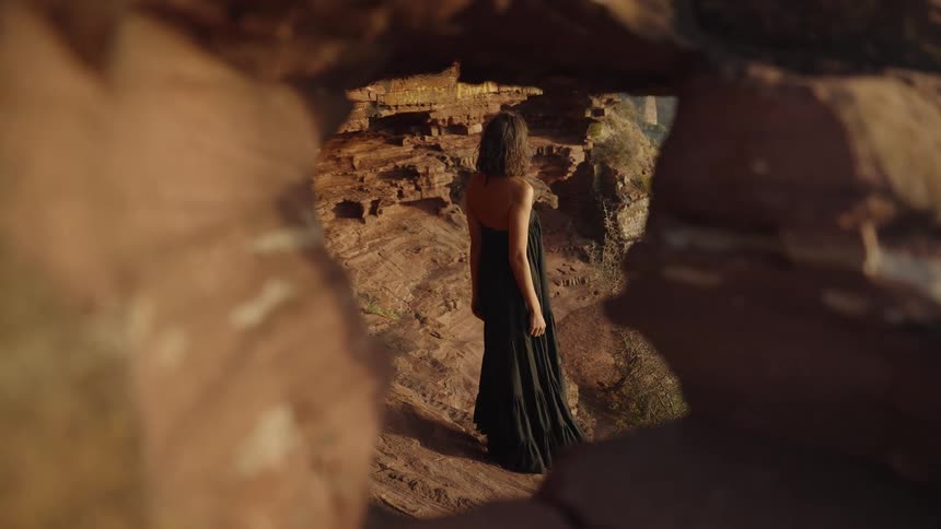 An elegant woman in a flowing black dress stands within a red rock canyon, framed by a natural stone arch at golden hour