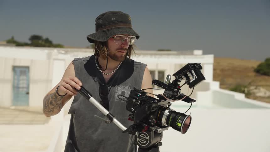 A tattooed videographer in a bucket hat and glasses operates a professional cinema camera on a tripod on a white rooftop