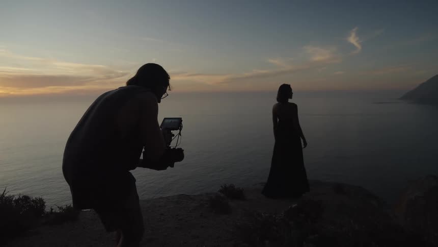 A videographer films a silhouetted model in a long dress as she poses on a cliff overlooking the ocean at sunset