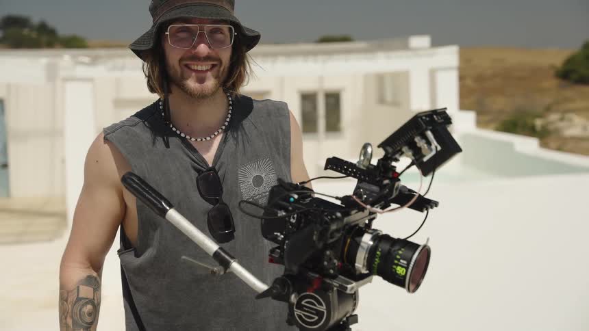 A young tattooed filmmaker in a bucket hat smiles behind a professional cinema camera on a bright, sunny day