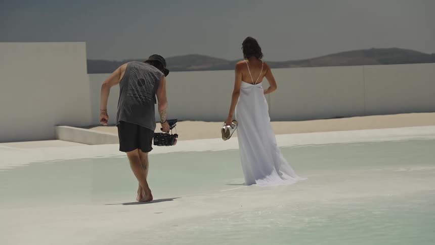 A cameraman follows a model in a flowing white gown as they both walk barefoot through a shallow pool during a photoshoot in slow motion