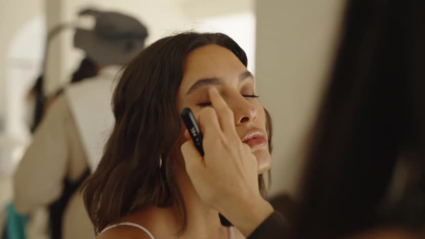 A makeup artist delicately applies eyeshadow with her finger to a model's eyelid in a backstage setting