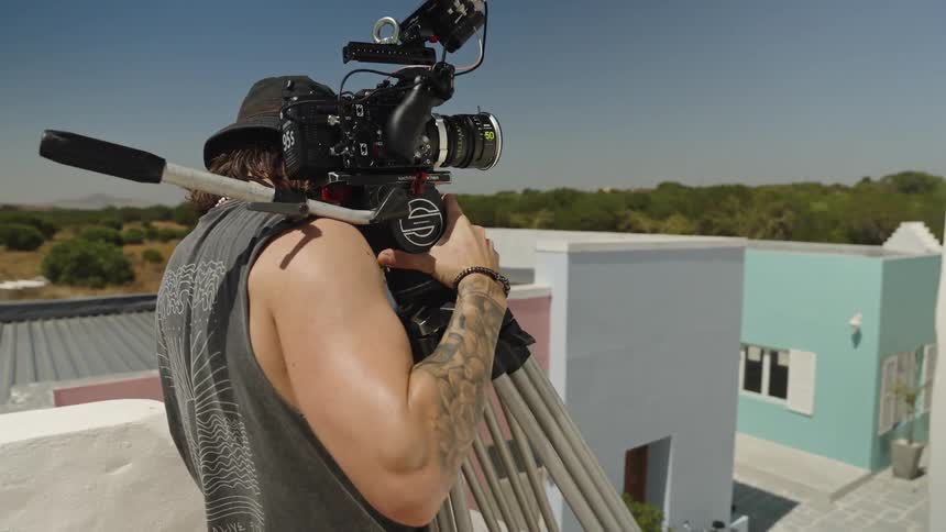 Behind-the-scenes shot following a cameraman as he operates a professional cinema camera on a tripod from a modern, colorful rooftop