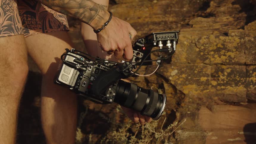 A filmmaker with tattoos adjusts their professional cinema camera rig while filming on location in nature