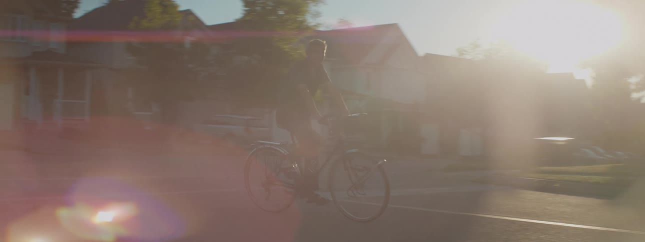 A man peacefully rides his bicycle through a quiet suburban neighborhood as the golden hour sun flares