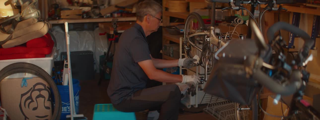 An older Asian man sits on a stool in his cluttered garage while carefully cleaning his bicycle
