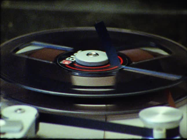 A grainy, vintage Super 8 close-up shot captures a spinning tape on a reel-to-reel recorder