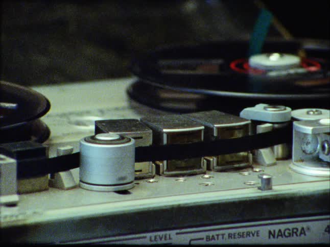 Grainy vintage film captures a close-up of a Nagra reel-to-reel tape recorder as it plays