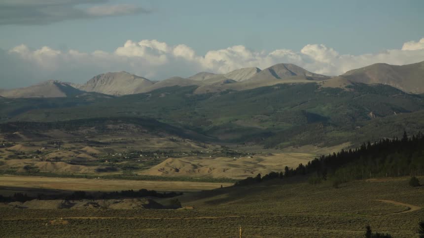 A tranquil and static wide shot shows a vast mountain valley and distant peaks under a blue sky with soft clouds