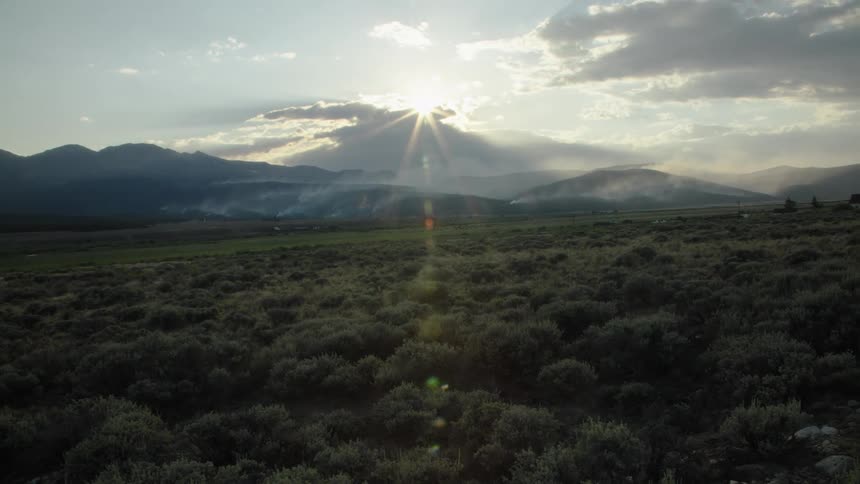 A slow zoom out reveals a scenic mountain valley with smoke rising from the foothills as the sun sets in the distance