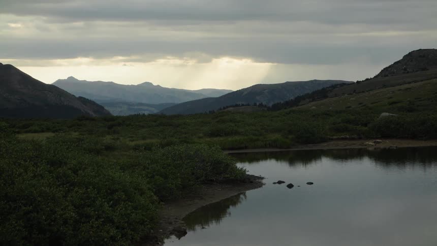 A calm mountain lake reflects the dramatic, cloudy sky as sunbeams break through over a distant mountain range