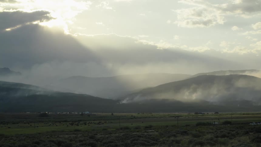 Ethereal sun rays shine through clouds onto a smoky, mountainous landscape during a hazy sunset