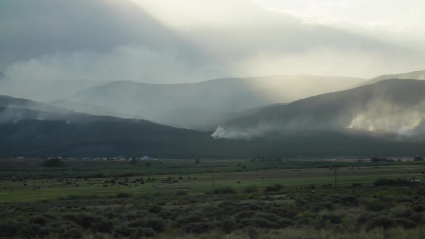 Smoky plumes from a distant wildfire rise over a vast mountain valley as sunbeams break through the clouds