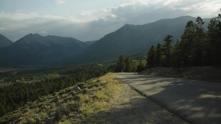A winding country road overlooks a vast, forested mountain valley under a cloudy sky