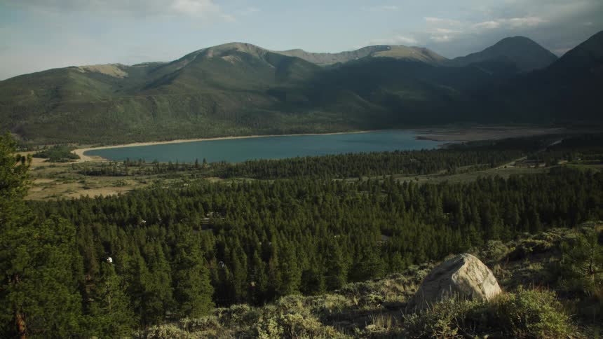A breathtaking wide-angle view of a serene mountain lake nestled in a valley surrounded by a dense evergreen forest and majestic peaks