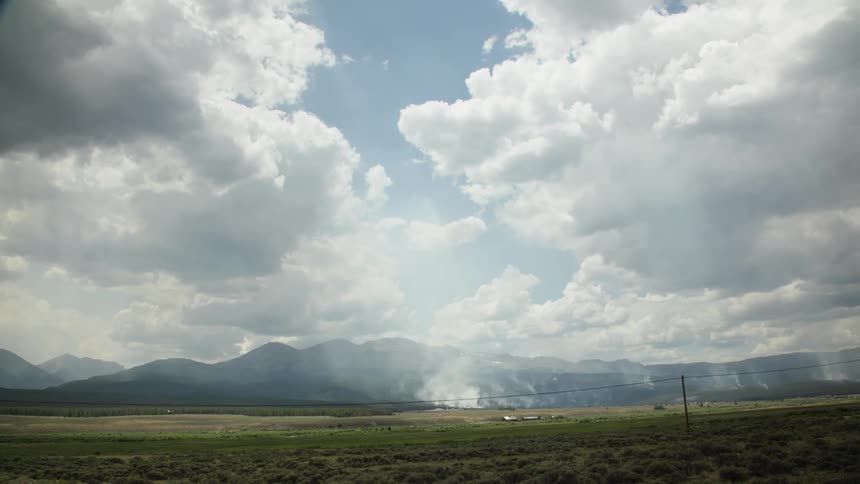 Smoke from a wildfire rises into the cloudy sky above a vast mountain landscape